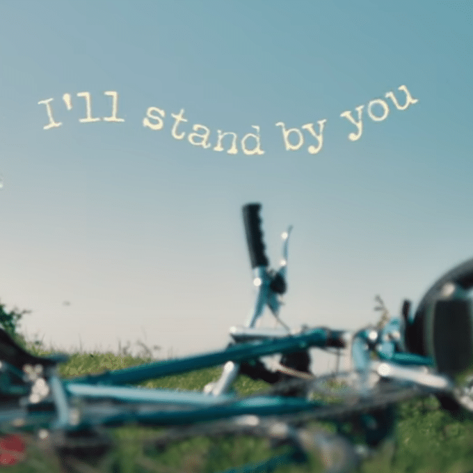La phrase "I'll Stand By You" est écrit au-dessus d'un vélo qui est posé dans l'herbe.