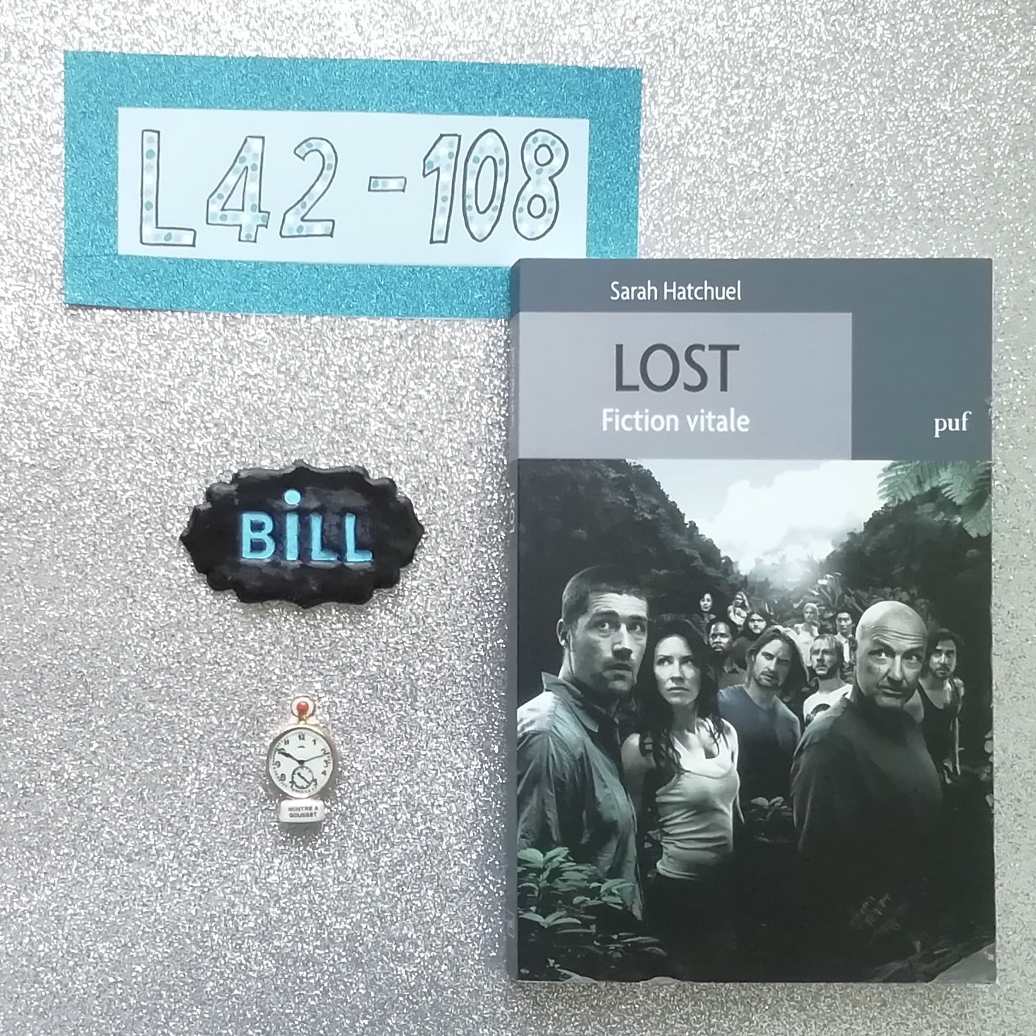 Le livre "Lost fiction vitale" se trouve à côté d'une fève qui représente une horloge. Au dessus, on retrouve également le prénom Bill réalisé en pâte polymer.